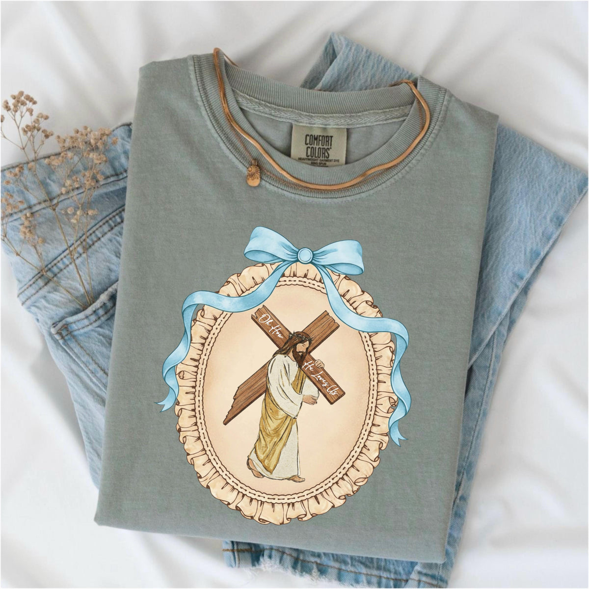 May include: Sage green Comfort Colors t-shirt with a graphic of Jesus carrying a cross, framed by a blue ribbon and a decorative oval. The text "Oh How He Loves Us" is visible on the cross. A brown leather necklace is draped on the shirt.
