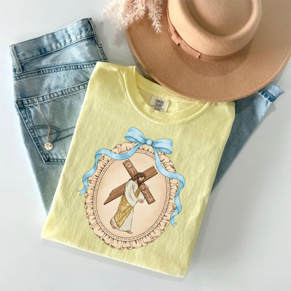 May include: A light yellow t-shirt featuring a religious illustration of Jesus carrying a cross, framed by a blue ribbon and a decorative border. The shirt is paired with light blue jeans and a tan hat, creating a casual, everyday look.