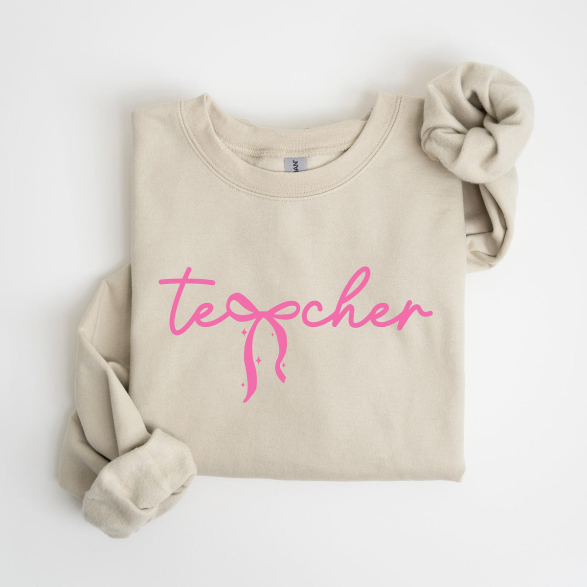 May include: A beige sweatshirt with the word "teacher" in pink cursive script. The word "teacher" has a pink bow design in the center. The sleeves are folded up, and a matching scrunchie is on the right side.