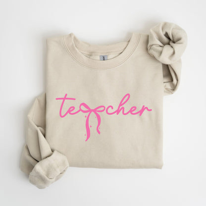 May include: A beige sweatshirt with the word "teacher" in pink cursive script. The word "teacher" has a pink bow design in the center. The sleeves are folded up, and a matching scrunchie is on the right side.