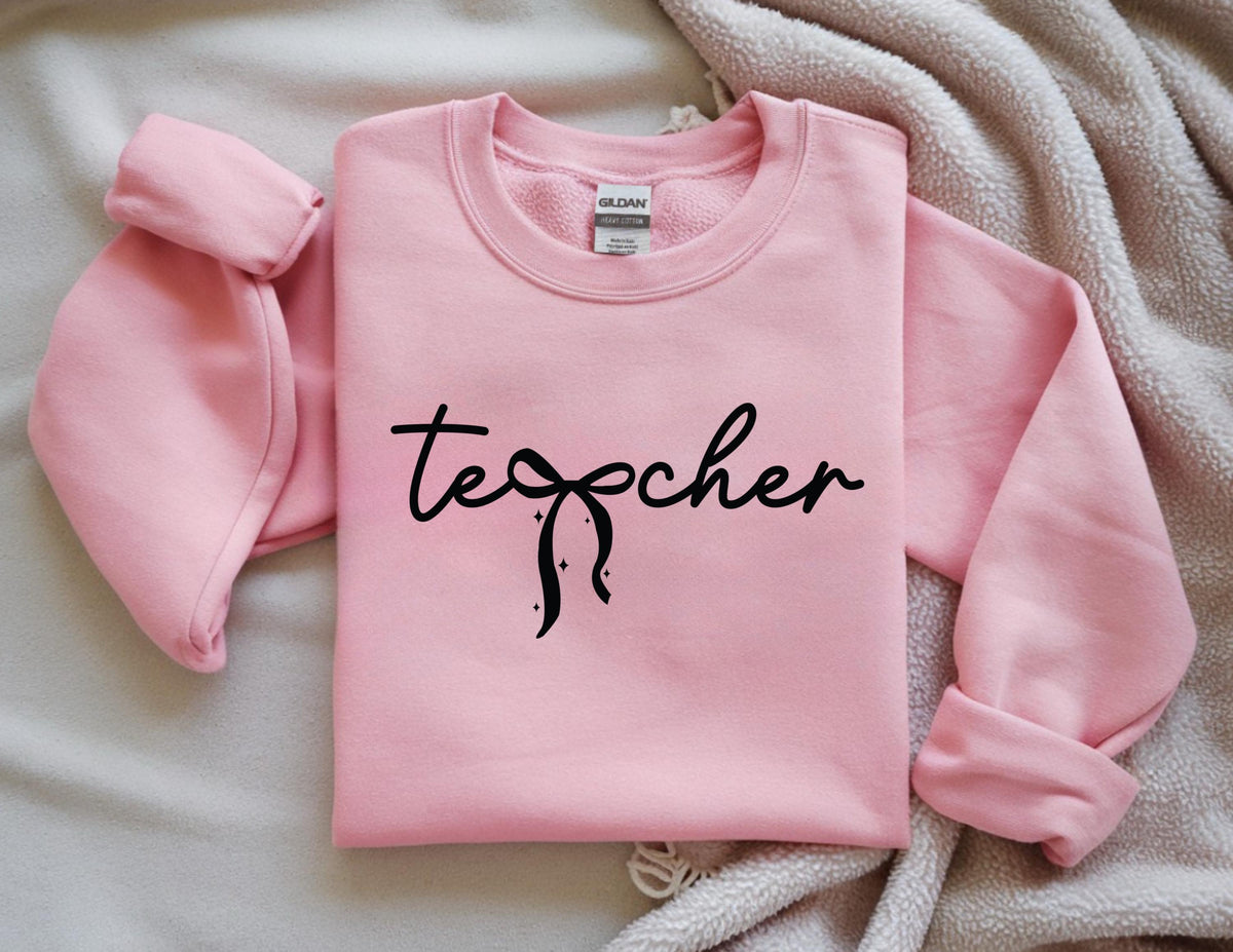 May include: A light pink sweatshirt with the word "teacher" in black script. The word features a bow design. The sleeves are rolled up. The sweatshirt is laying on a light brown blanket.