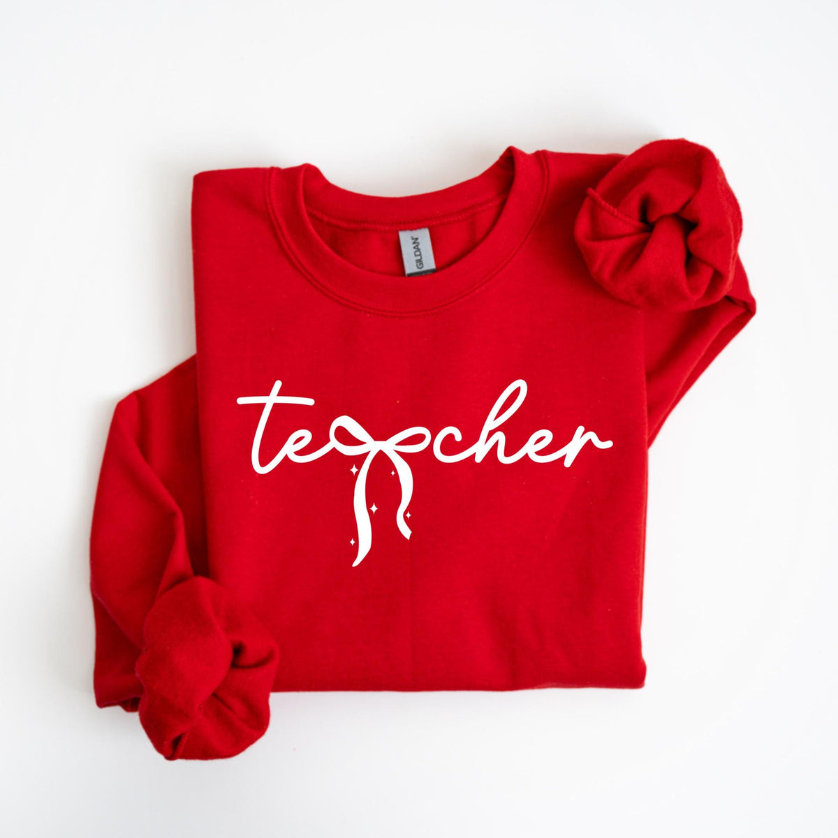 May include: A red sweatshirt with the word "teacher" in white script, featuring a bow design. The long sleeves are folded. The Gildan brand label is visible on the inside collar.