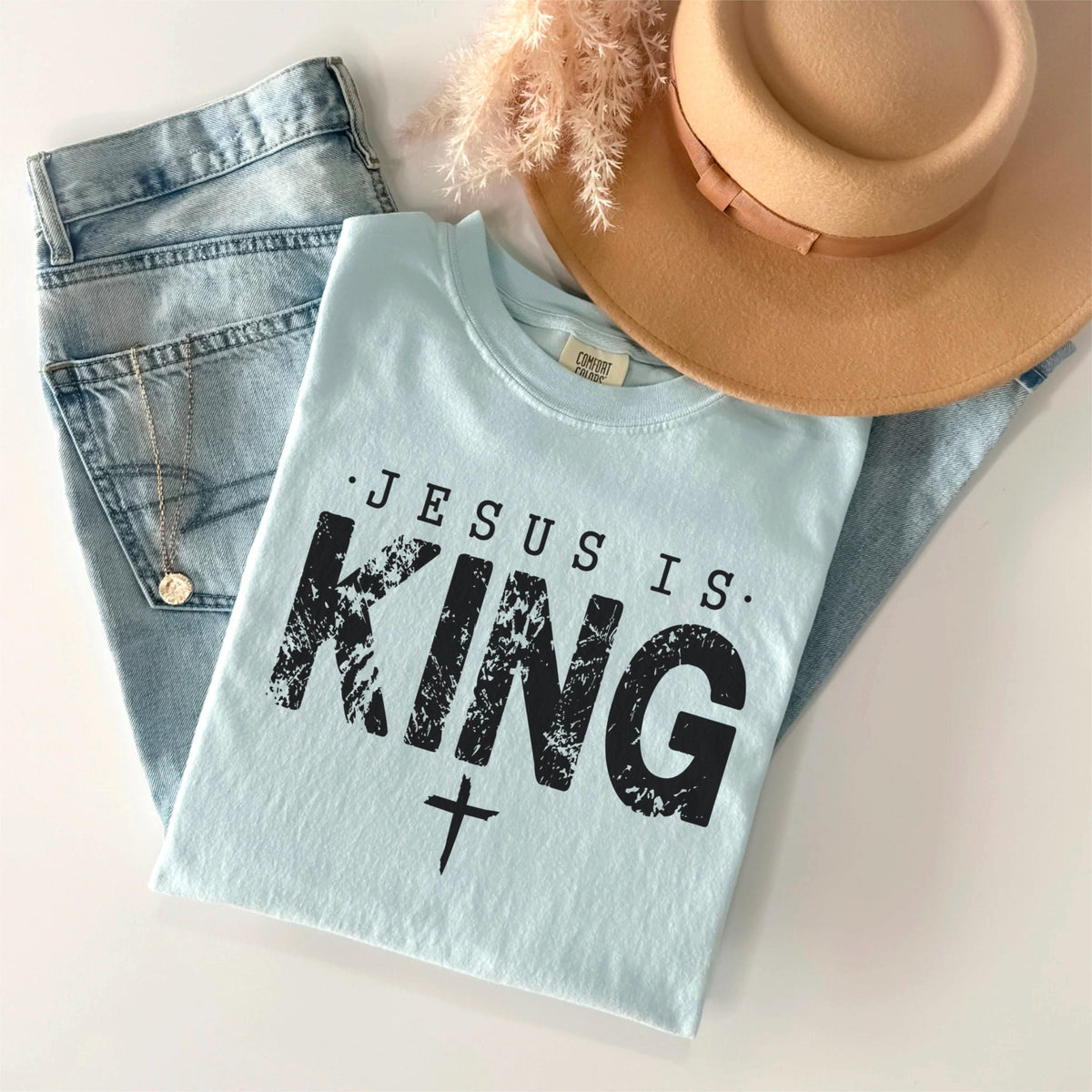 May include: A light blue t-shirt with the text "JESUS IS KING" in black, along with a cross graphic. The shirt is paired with light wash denim jeans, a tan hat, and a gold necklace. A sprig of dried flowers is also present.