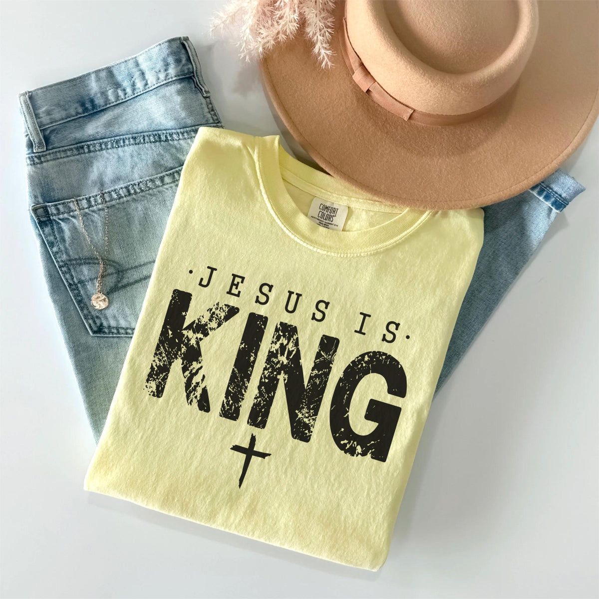 May include: A light yellow t-shirt with the text "JESUS IS KING" in black, along with a cross graphic. The shirt is paired with light blue jeans and a tan hat. A necklace is visible in the jeans pocket.