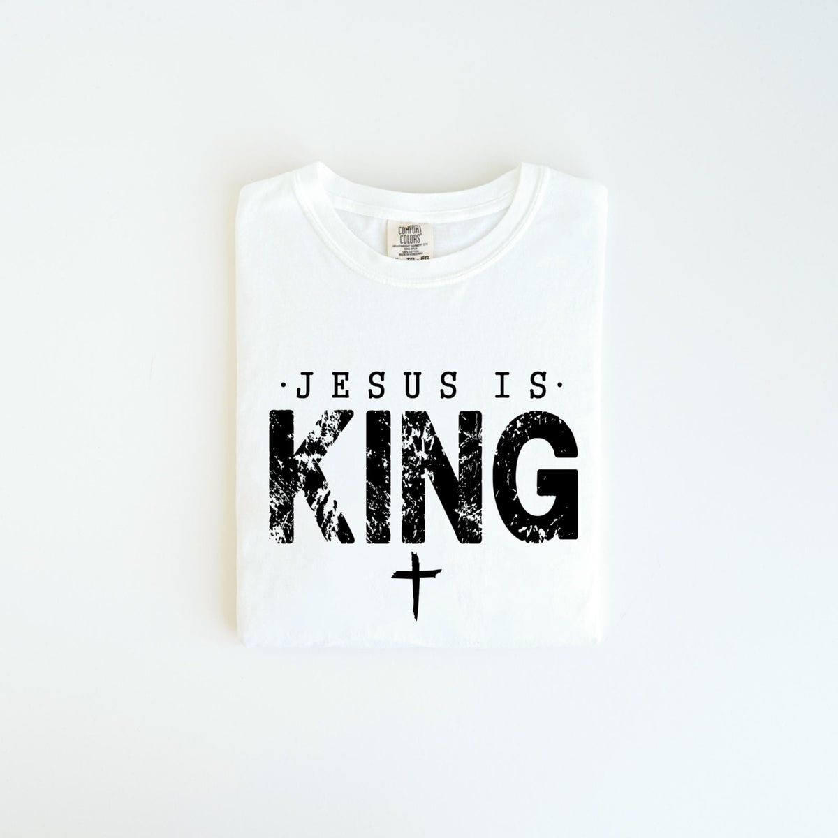 May include: White t-shirt with the text "JESUS IS KING" in black, distressed font. A small cross is below the word "KING". The shirt is folded neatly on a white surface.