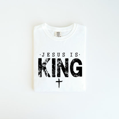 May include: White t-shirt with the text "JESUS IS KING" in black, distressed font. A small cross is below the word "KING". The shirt is folded neatly on a white surface.