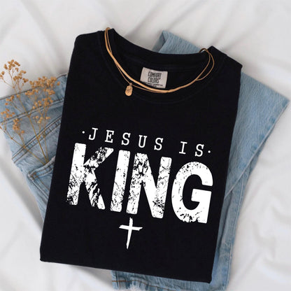 May include: Black t-shirt with the text "JESUS IS KING" in distressed white lettering and a white cross. The shirt is folded on top of blue denim jeans. A gold necklace rests on the shirt.