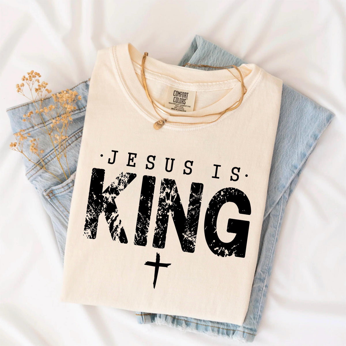 May include: Cream-colored t-shirt with the text "JESUS IS KING" in black, distressed font, and a cross below. The shirt is folded on top of blue denim jeans, with a gold necklace and dried flowers. The shirt is from Comfort Colors.