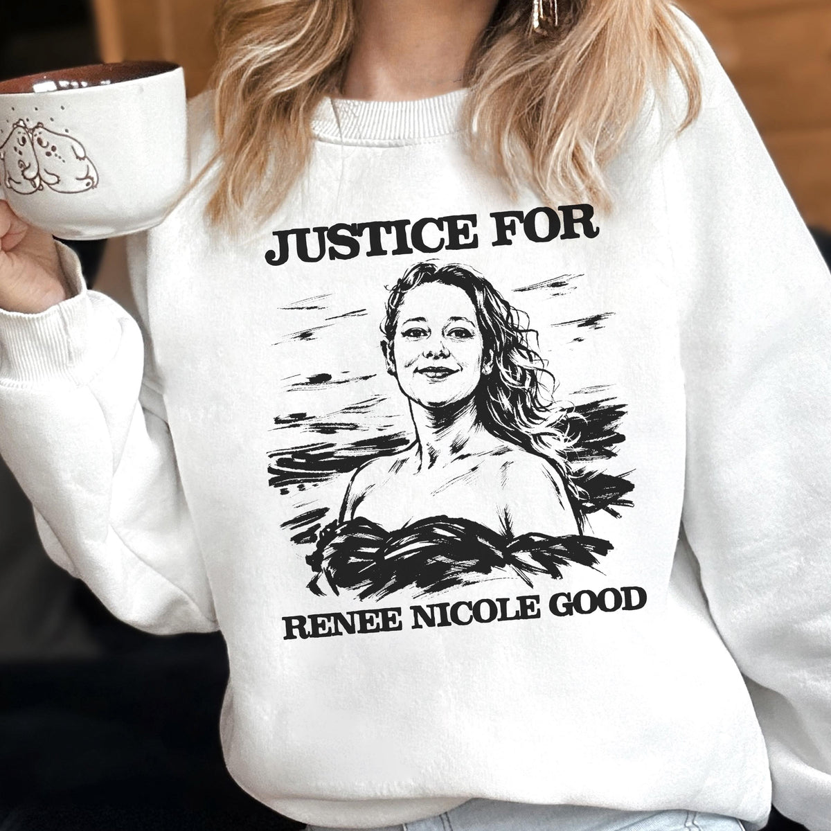 May include: White sweatshirt with the text "JUSTICE FOR" above a black and white illustration of a woman, and the text "RENEE NICOLE GOOD" below. The person is holding a white mug with a brown interior and a cartoon drawing.