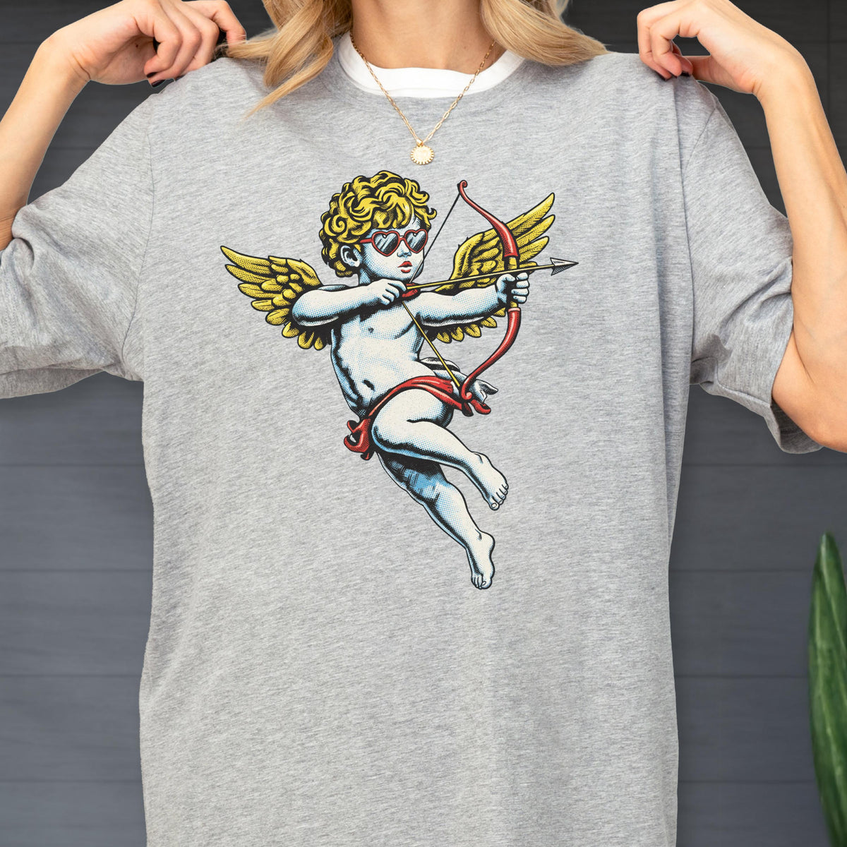 May include: A heather gray short-sleeve t-shirt featuring a graphic print of a cherub with curly blonde hair, wearing red heart-shaped sunglasses, and holding a red bow and arrow with yellow wings. The design is centered on the chest area.