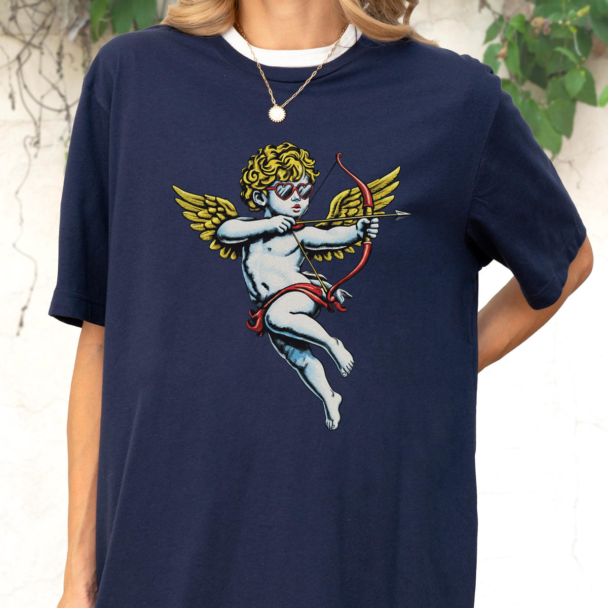 May include: A navy blue short-sleeve t-shirt featuring a graphic print of a cherub with golden wings, wearing heart-shaped sunglasses and aiming a red bow and arrow. The cherub has curly blonde hair and is depicted in a classic artistic style.