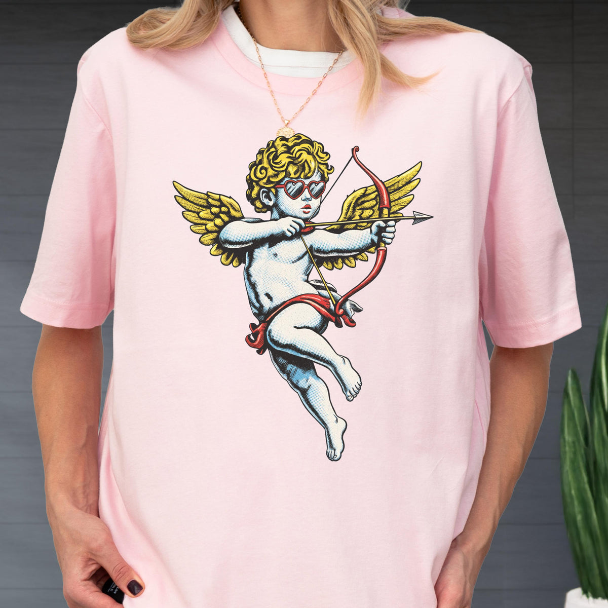 May include: A light pink short-sleeve t-shirt featuring a central graphic of a cherub with golden wings, wearing red heart-shaped sunglasses and aiming a red bow and arrow. The cherub has curly blonde hair and is depicted in a classic art style.