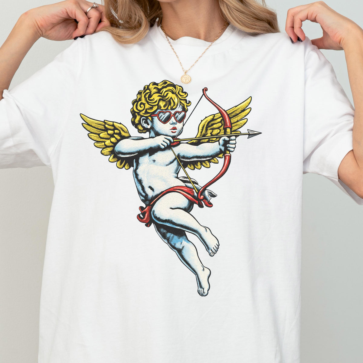 May include: A white t-shirt featuring a graphic print of a cherub with curly blonde hair, wearing red heart-shaped sunglasses, and holding a red bow and arrow. The cherub has large yellow wings and is depicted in a classic flying pose.
