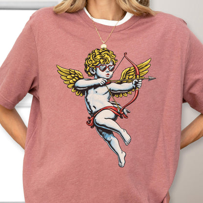 May include: A dusty rose short-sleeve t-shirt featuring a graphic print of a cherub with curly blonde hair, wearing red heart-shaped sunglasses, and holding a red bow and arrow. The cherub has large yellow wings and is depicted in a classic style.