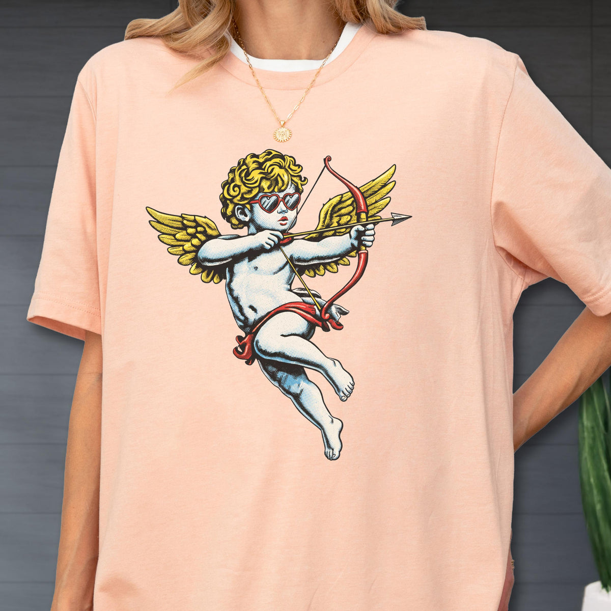 May include: A peach-colored t-shirt featuring a graphic print of a cherub with curly blonde hair, wearing heart-shaped sunglasses, and holding a red bow and arrow. The cherub has large yellow wings and is depicted in a classic flying pose.