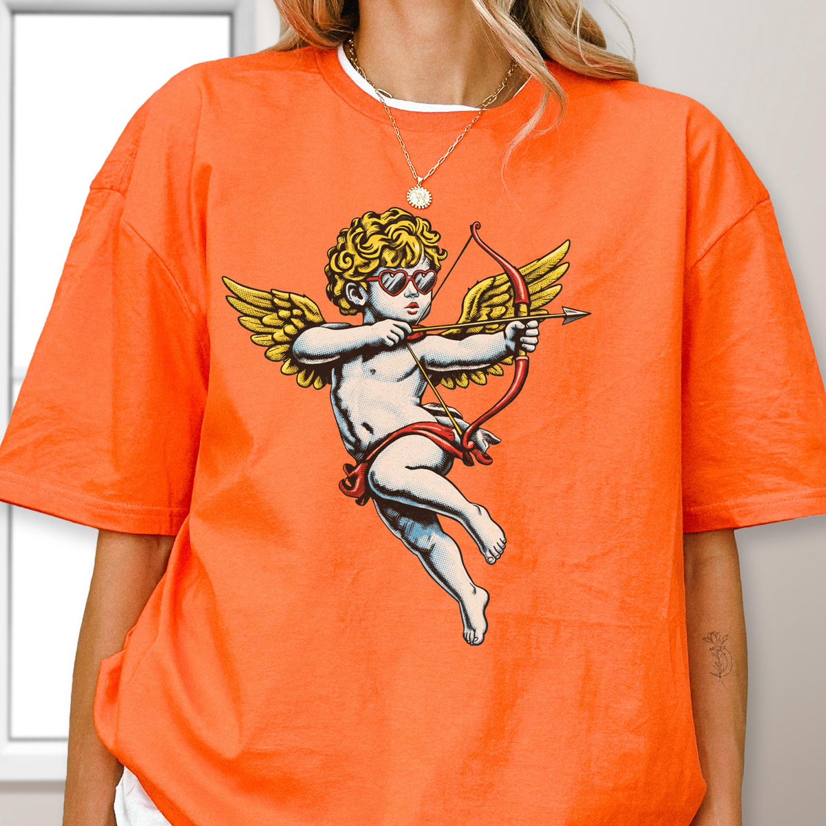 May include: A bright orange t-shirt featuring a graphic print of a cherub with golden wings, curly hair, and red heart-shaped sunglasses. The cherub is depicted in a flying pose, holding a red bow and arrow, rendered in a classic illustrative style.