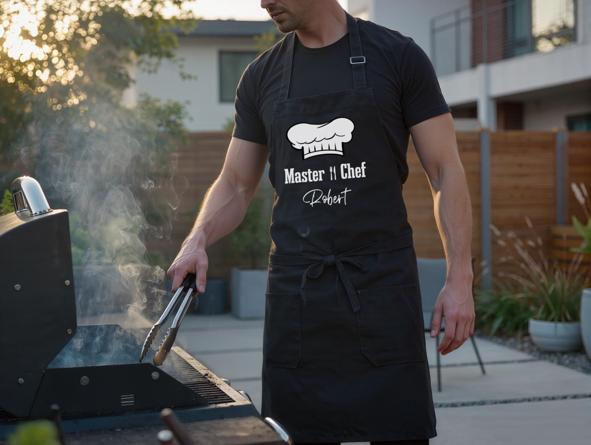 May include: A black apron with the text "Master Chef Robert" and a chef's hat logo. The apron is worn by a person using tongs to cook on a black grill, with smoke rising. The scene takes place outdoors.