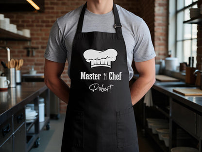 May include: Black apron with a white chef's hat graphic and the text "Master Chef Robert". The apron has two front pockets and adjustable neck straps. The person is wearing a gray t-shirt.