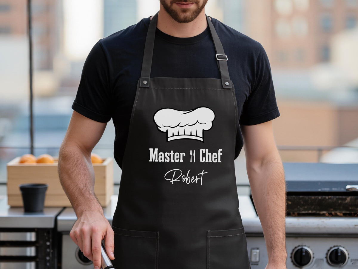 May include: Black apron with a white chef's hat graphic and the text "Master Chef" and the name "Robert". The apron has two front pockets and gray straps. The person is wearing a black t-shirt.