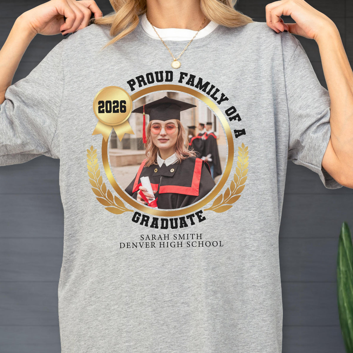 May include: A light gray t-shirt featuring a graduation design. The design includes a photo of a graduate in a cap and gown, surrounded by a gold laurel wreath and the text "PROUD FAMILY OF A GRADUATE" and "2026". The graduate's name and school are also printed.