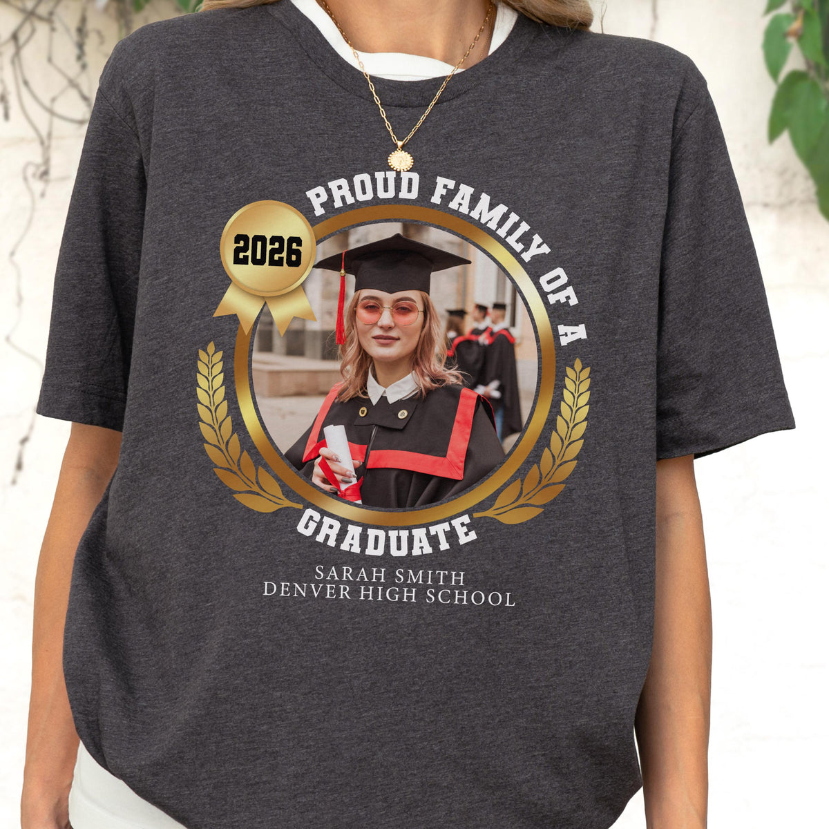 May include: Dark gray t-shirt featuring a graduation design. The design includes a photo of a graduate in a cap and gown, surrounded by text that reads "Proud Family of a Graduate" and the year "2026". The graduate's name and high school are also displayed.