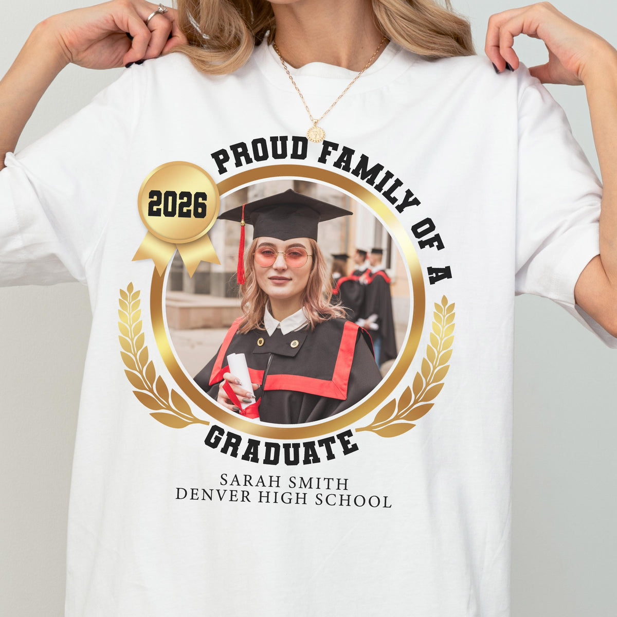 May include: White t-shirt featuring a graduation design. The design includes a photo of a graduate in a cap and gown, surrounded by a gold laurel wreath. Text reads "Proud Family of a Graduate" and "2026", along with the graduate's name and high school.
