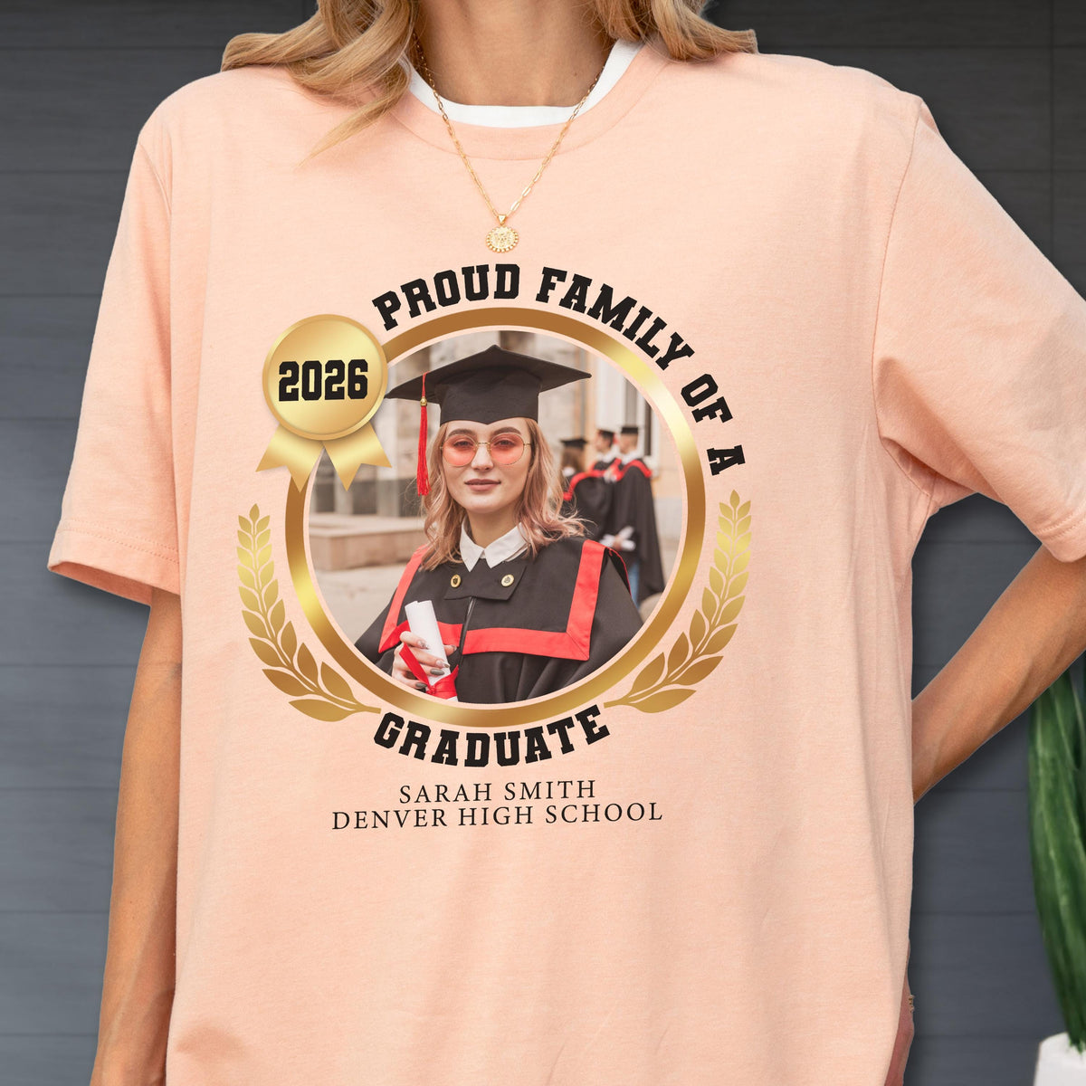 May include: A peach-colored t-shirt featuring a gold-colored circular design with the text "PROUD FAMILY OF A GRADUATE" and "2026". The design includes a photo of a graduate in a cap and gown, with the name "SARAH SMITH DENVER HIGH SCHOOL" below.