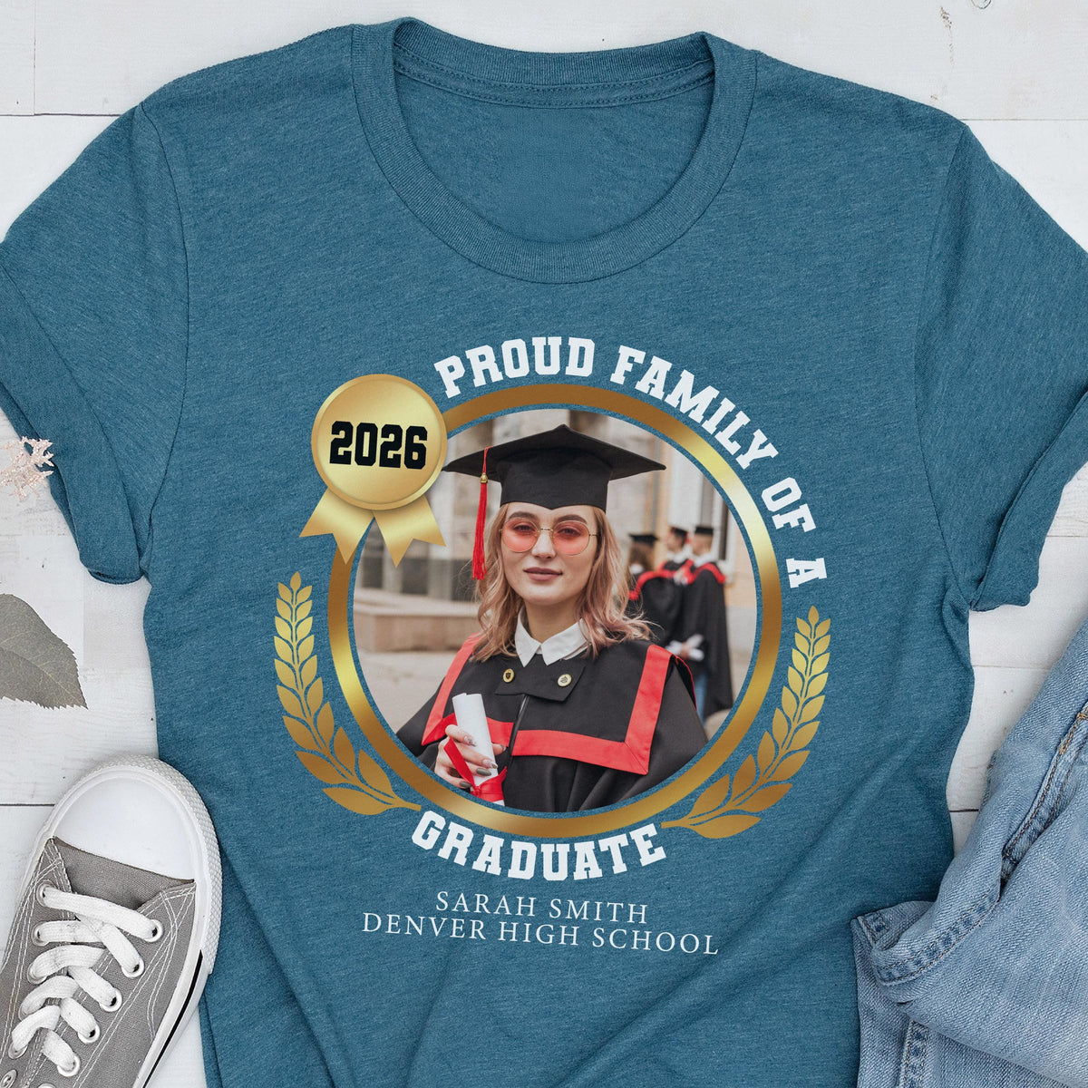 May include: Teal t-shirt featuring a graduation design. The design includes a photo of a graduate in a cap and gown, surrounded by a gold laurel wreath and the text "Proud Family of a Graduate" and "2026". The graduate's name and high school are also printed.