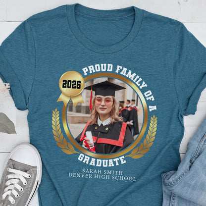 May include: Teal t-shirt featuring a graduation design. The design includes a photo of a graduate in a cap and gown, surrounded by a gold laurel wreath and the text "Proud Family of a Graduate" and "2026". The graduate's name and high school are also printed.