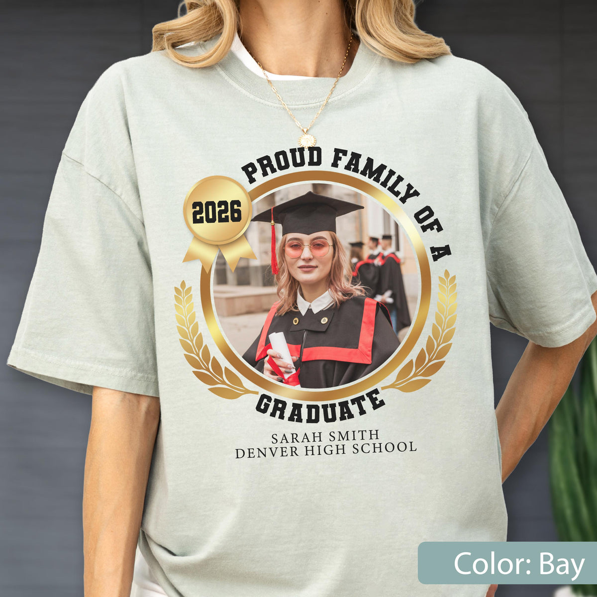 May include: Bay-colored t-shirt with a graduation-themed design. The design includes a photo of a graduate in a cap and gown, surrounded by text that reads "Proud Family of a Graduate 2026" and the graduate's name and school.
