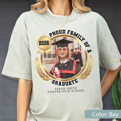 May include: Bay-colored t-shirt with a graduation-themed design. The design includes a photo of a graduate in a cap and gown, surrounded by text that reads "Proud Family of a Graduate 2026" and the graduate's name and school.
