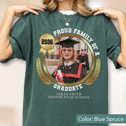 May include: Blue spruce t-shirt with a gold emblem featuring a graduate's photo, the text "Proud Family of a Graduate," and the year 2026. The graduate is wearing a cap and gown. The shirt also includes the graduate's name and high school.