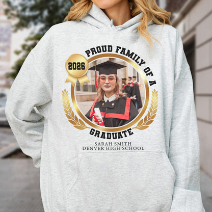 May include: Light gray hoodie with a circular graphic. The graphic features a photo of a graduate in a cap and gown, surrounded by text that reads "Proud Family of a Graduate" and "2026". The graduate's name and school are also displayed.