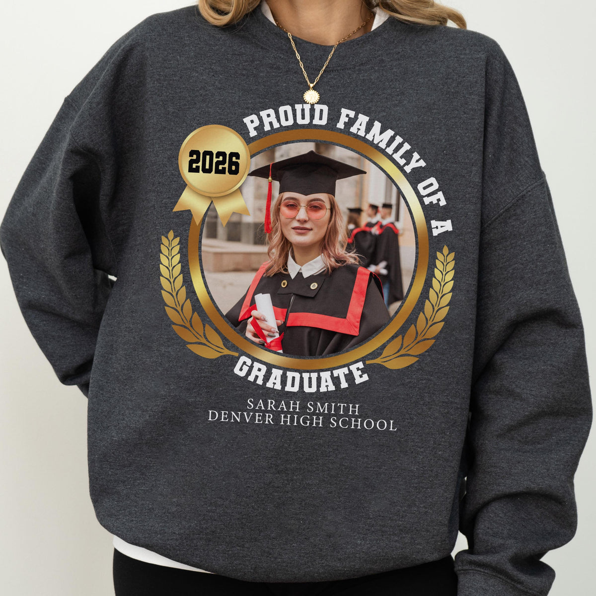 May include: Dark gray sweatshirt with a gold emblem featuring a graduate's photo, the text "Proud Family of a Graduate," and "2026." The graduate's name and high school are printed below. A gold necklace is visible.