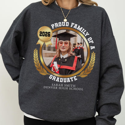May include: Dark gray sweatshirt with a gold emblem featuring a graduate's photo, the text "Proud Family of a Graduate," and "2026." The graduate's name and high school are printed below. A gold necklace is visible.
