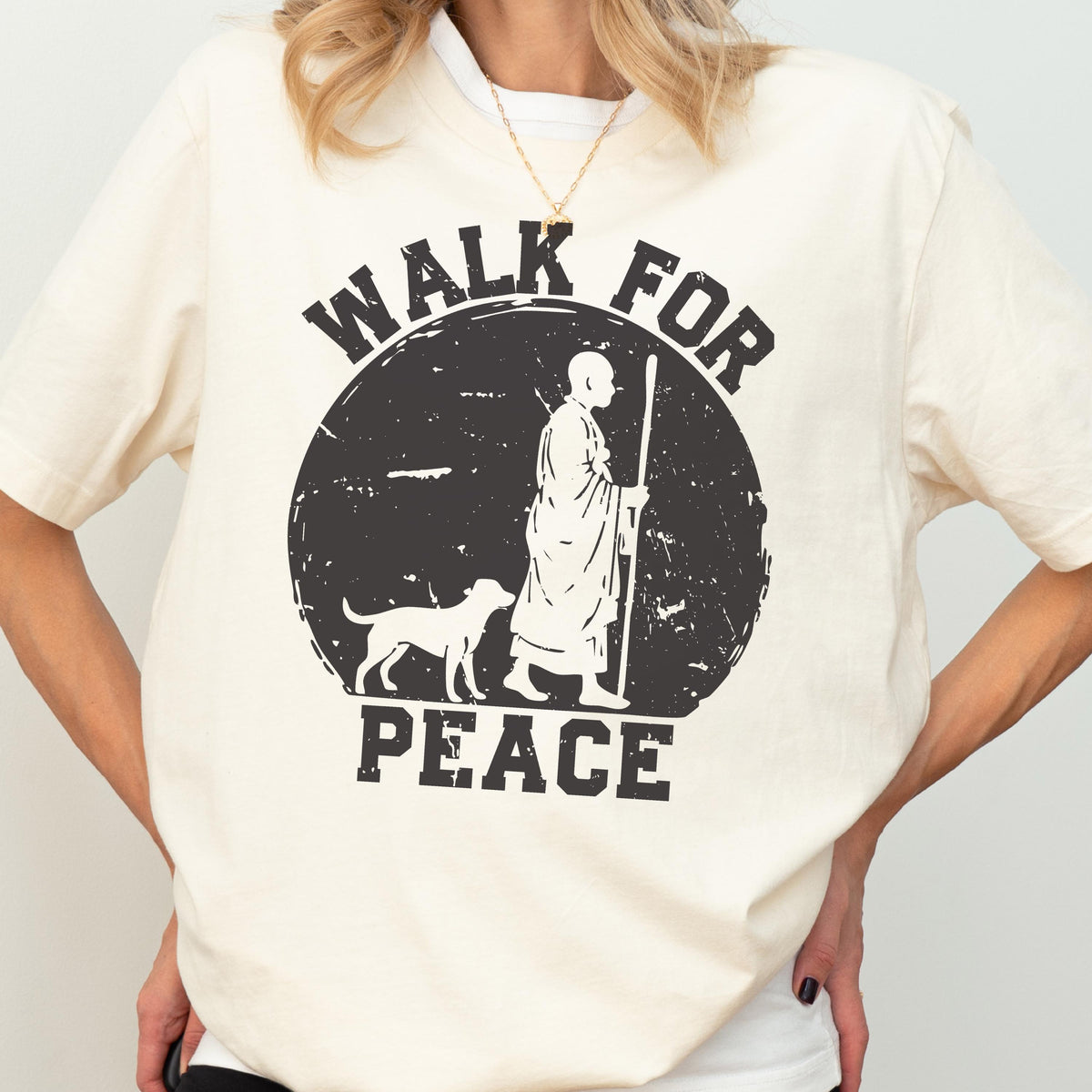May include: A cream-colored short-sleeve t-shirt featuring a black graphic of a person in robes walking with a dog and a staff inside a circle. The text "WALK FOR PEACE" is printed in a distressed black font above and below the circular graphic.