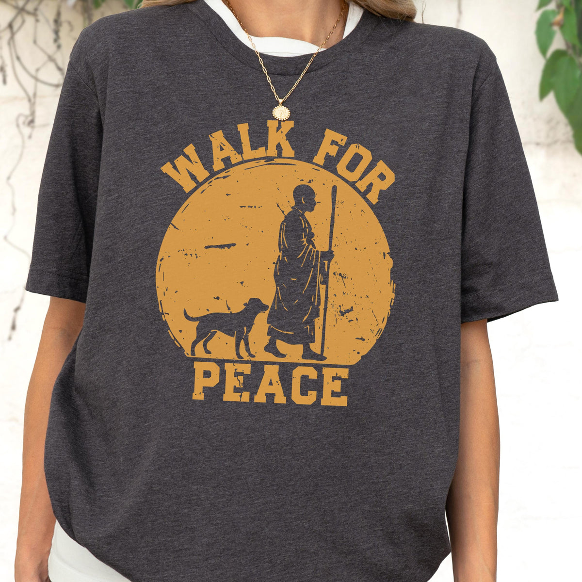 May include: A dark charcoal gray short-sleeve t-shirt featuring a distressed mustard yellow graphic of a person walking with a dog and a staff inside a circle. The text "WALK FOR PEACE" is printed in a bold, blocky font above and below the circle.