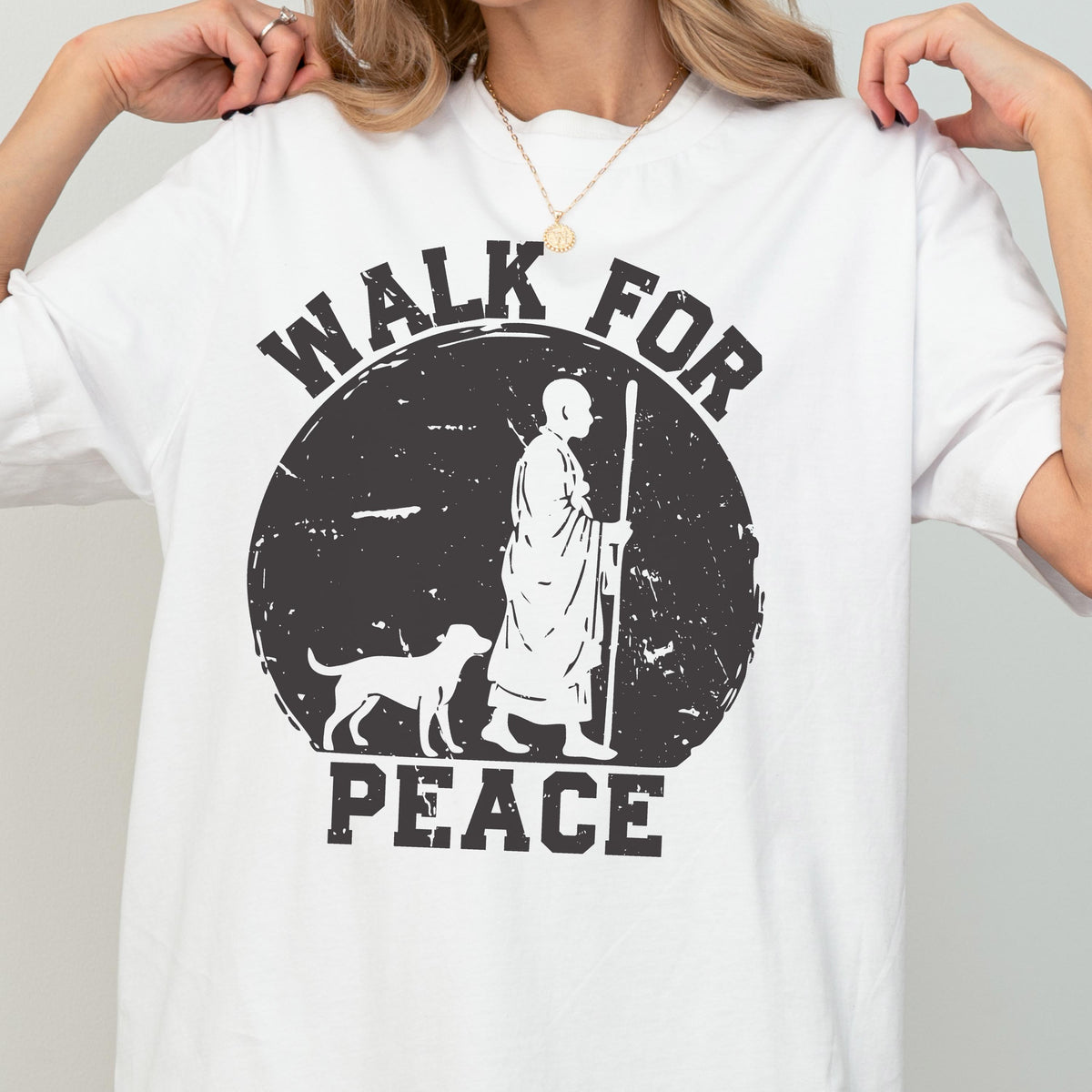 May include: A white short-sleeved t-shirt featuring a black graphic of a monk walking with a dog inside a distressed circle. The text "WALK FOR PEACE" is printed in black block letters above and below the circular graphic design.