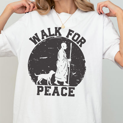 May include: A white short-sleeved t-shirt featuring a black graphic of a monk walking with a dog inside a distressed circle. The text "WALK FOR PEACE" is printed in black block letters above and below the circular graphic design.