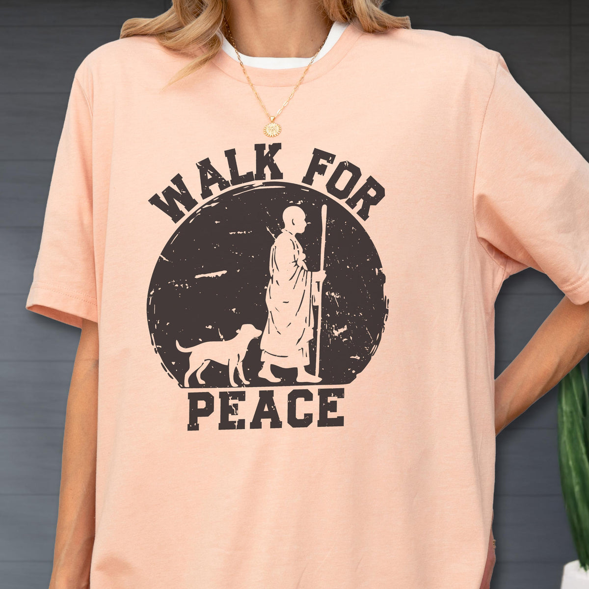 May include: A peach-colored short-sleeve t-shirt featuring a black circular graphic with a distressed texture. The design depicts a person in robes holding a staff walking with a dog, accompanied by the text WALK FOR PEACE in bold lettering.