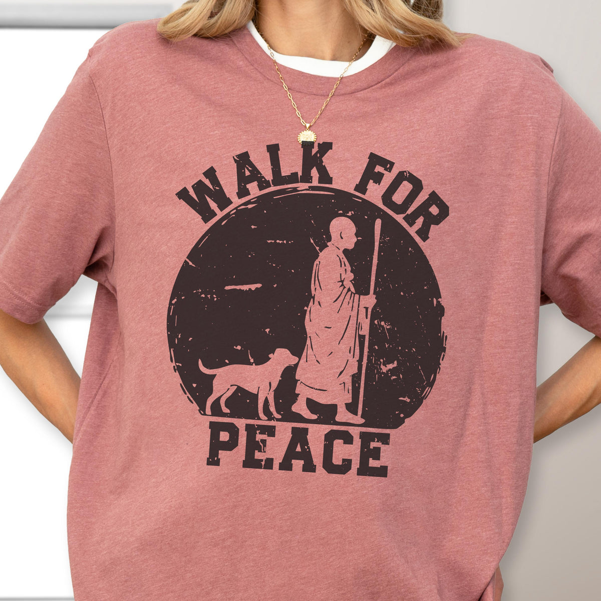 May include: A dusty rose short-sleeve t-shirt featuring a black circular graphic. The design depicts a person in robes holding a staff walking with a dog, surrounded by the text WALK FOR PEACE in a distressed, bold, block-style font.