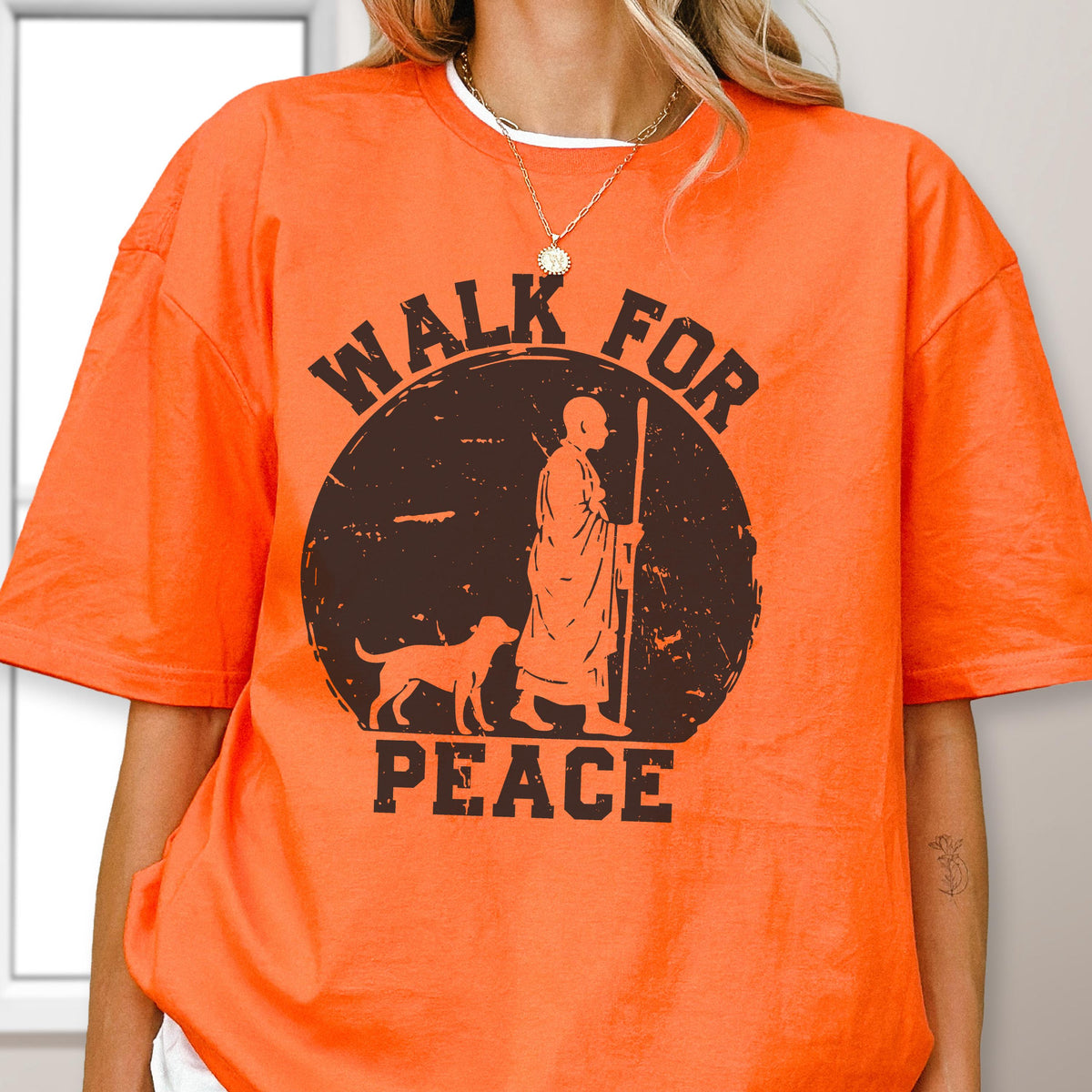 May include: A bright orange short-sleeved t-shirt featuring a dark brown circular graphic. The design depicts a person in robes walking with a staff alongside a dog, with the text "WALK FOR PEACE" printed in bold, distressed capital letters.