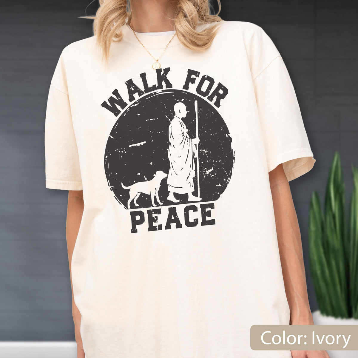 May include: Ivory-colored t-shirt featuring a distressed black graphic. The graphic depicts a figure with a staff, a dog, and the words "WALK FOR PEACE" in a circular design. The shirt has short sleeves and a relaxed fit.