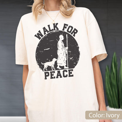 May include: Ivory-colored t-shirt featuring a distressed black graphic. The graphic depicts a figure with a staff, a dog, and the words "WALK FOR PEACE" in a circular design. The shirt has short sleeves and a relaxed fit.
