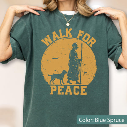 May include: Blue spruce t-shirt with a distressed graphic. The design features a figure with a dog walking in front of a yellow sun, with the words "WALK FOR PEACE" in a circular layout.