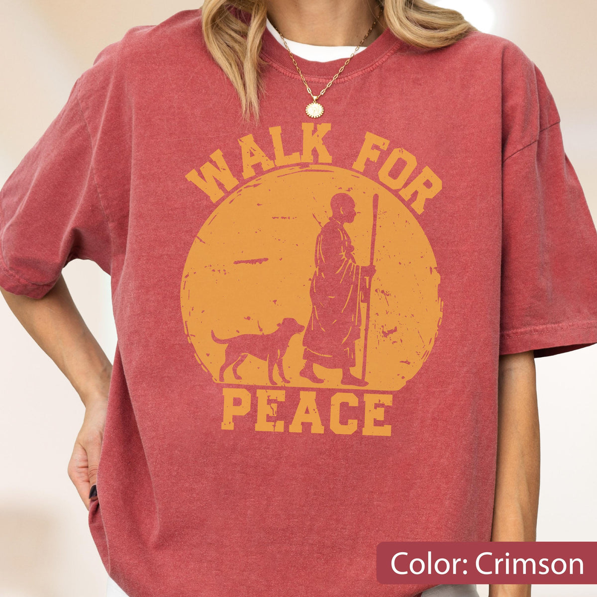 May include: Crimson-colored t-shirt with a graphic design. The design features a figure with a dog, a staff, and the words "WALK FOR PEACE" in an orange circle. The shirt has short sleeves and a crew neck.