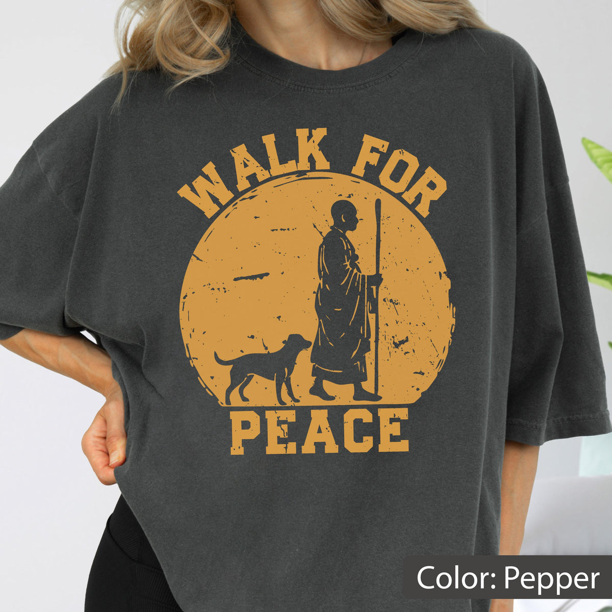 May include: Pepper-colored t-shirt with a distressed orange graphic. The graphic features a silhouetted figure with a staff, a dog, and the words "WALK FOR PEACE". The shirt is a relaxed fit.