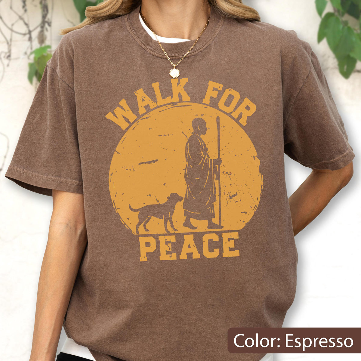 May include: Espresso-colored t-shirt featuring a distressed yellow graphic of a person with a dog, staff in hand, inside a circle. The words "WALK FOR PEACE" are printed above and below the image. The shirt is a solid color.
