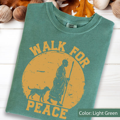 May include: Light green t-shirt with the words "WALK FOR PEACE" in orange, featuring a graphic of a person with a dog walking with a staff. The shirt has a crew neck and a tag that says "Comfort Colors".