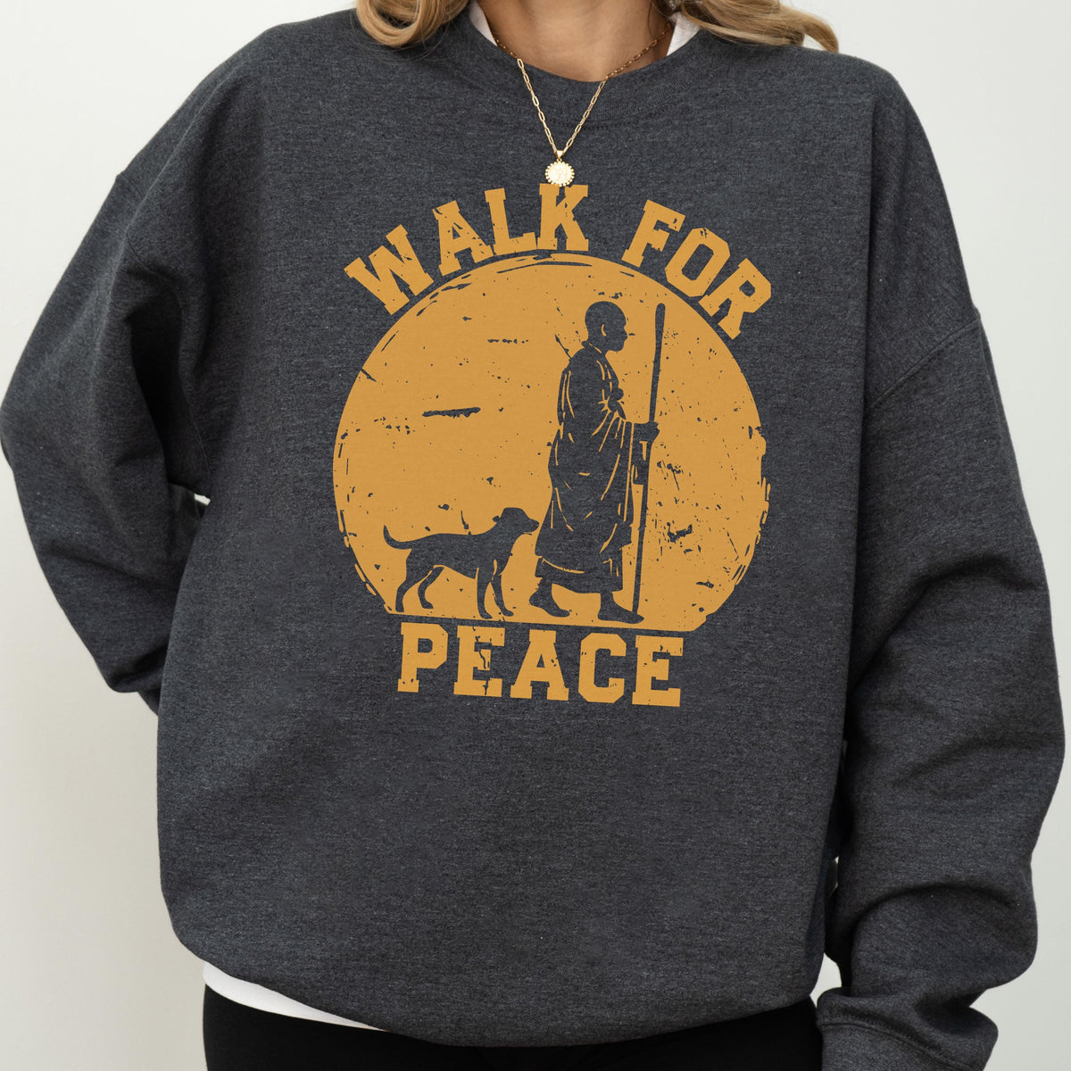 May include: Dark gray sweatshirt with a distressed orange graphic. The graphic features a person with a staff and a dog walking in front of a yellow sun with the words "WALK FOR PEACE". The sweatshirt has a crew neck and long sleeves.